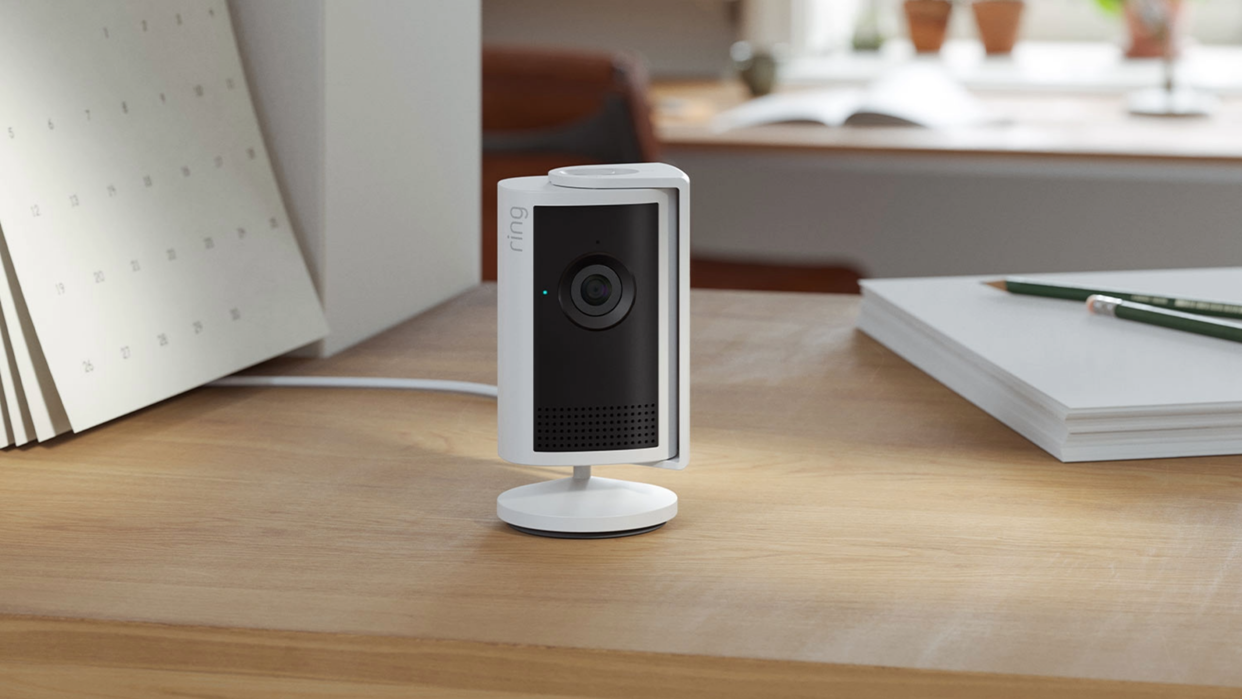 Ring Indoor Cam Plus in white on a home office desk with privacy cover on, surrounded by notebooks, plants, and natural light.